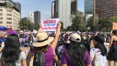 People walking along a street between high rise buildings. One is holding a sign 'Mexico es un Pais Feminicida' above her head. 