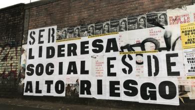 Poster with white background and bold black text 'SER LIDERESA SOCIAL ESDE ALTO RIESGO' attached to a high brick wall