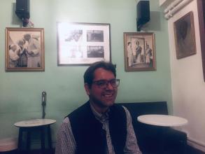 Adnan has short dark brown hair, is wearing glasses and smiling broadly at the camera. He's wearing a blue and white gingham shirt under a navy blue woollen waist coat. Behind him is a pale green painted wall with framed black and white photos of people.   