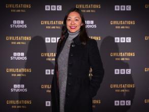 Jessica has long dark hair and is smiling at the camera. She's wearing a grey dress under a long black coat. She's standing against a 'BBC Civilisations: Rise and Fall' backdrop