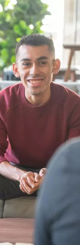 A male student wearing a red jumper sitting on a sofa in a shared lounge area. He is talking to two other students with their backs to the camera. 
