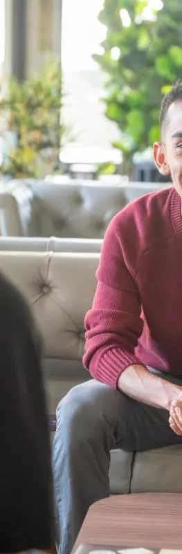 A male student wearing a red jumper sitting on a sofa in a shared lounge area. He is talking to two other students with their backs to the camera. 
