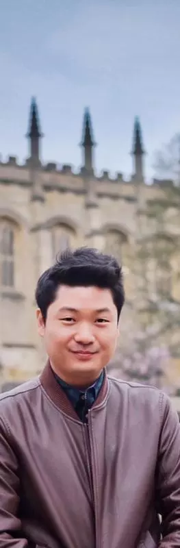 A man with dark hair and wearing a brown jacket over a blue shirt is standing in an Oxford college garden with a church behind him. 